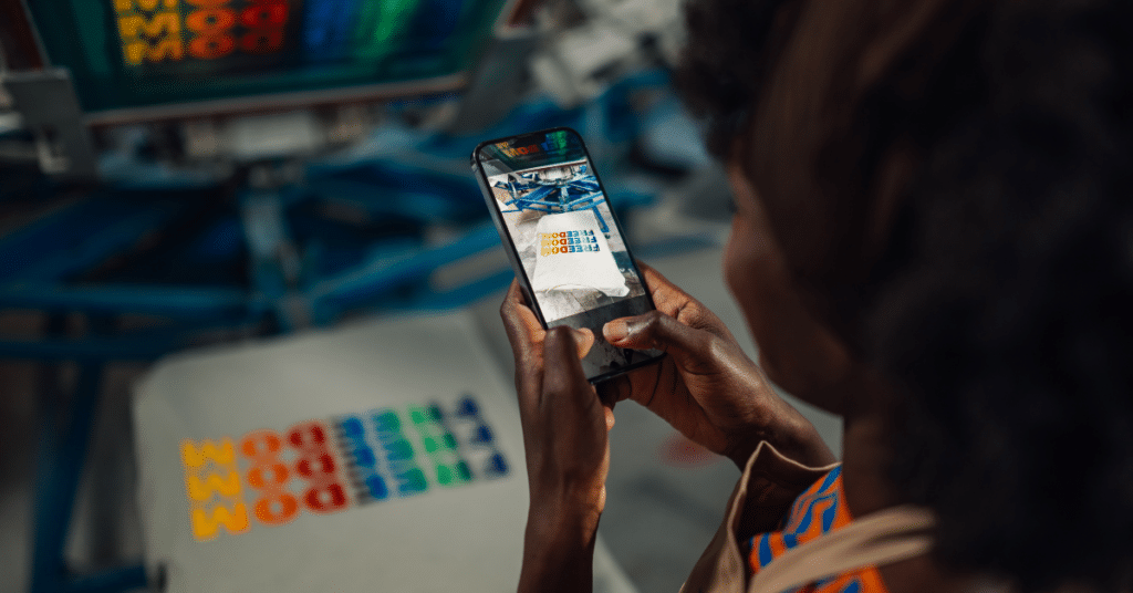 African woman using smartphone to document screen-printed t-shirts with bold messages, inside a modern local apparel production facility