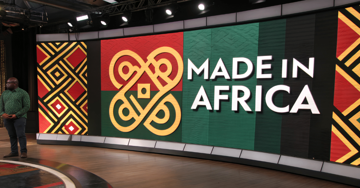 African presenter standing beside large Made in Africa studio display with bold traditional patterns and Pan-African colour scheme, highlighting African-made branding and cultural pride