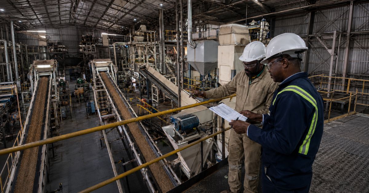 Two industrial supervisors wearing hard hats review documents inside a processing facility, representing operational continuity, industrial performance recalibration and resilience under pressure.